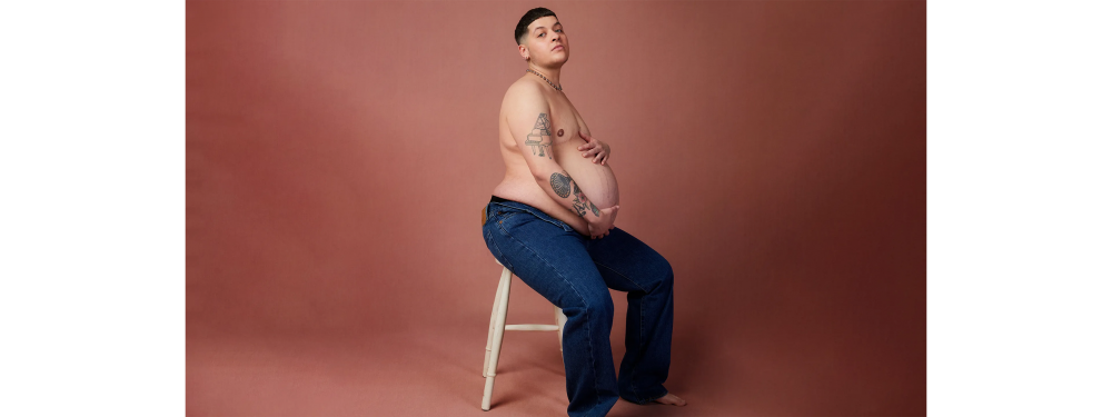 Logan Brown with their top off, exposing their pregnant belly
