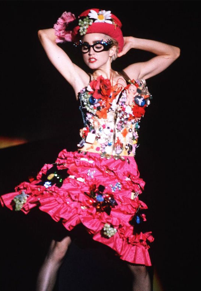 A photo of Madonna wearing a very camp dress on stage whilst performing. The dress is hot pink and decorated with lots of glittery objects, metallic flowers, sequins and miscellaneous objects like miniature dolls.