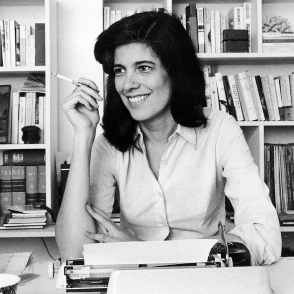 A photo of Susan Sontag wearing a white shirt at a desk, and smoking a cigarette.