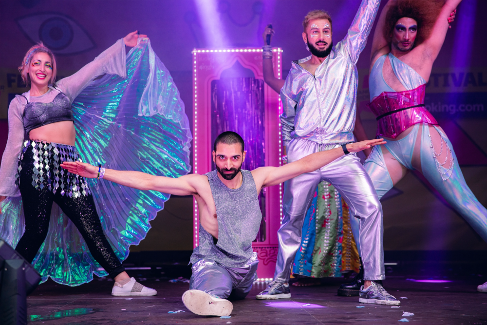 House of Spice perform on the Alan Turing Stage at the Gay Village Party wearing fabulous metallic outfits. One person poses in the splits whilst another three performers lunge and smile at the camera.