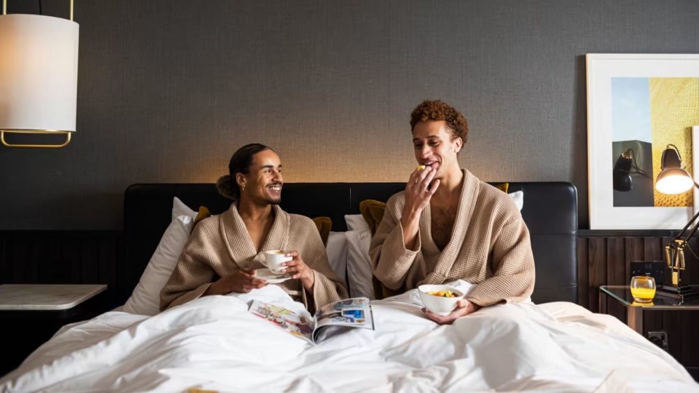 Couple in hotel robes holding coffee cups and laughing together, in a hotel bed..
