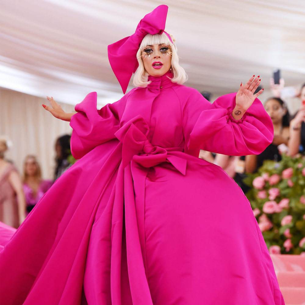 A photo of Lady Gaga wearing a large, very camp hot pink dress, decorated with large bows. She has bleach blonde hair and wears a large hot pink bow on her head.
