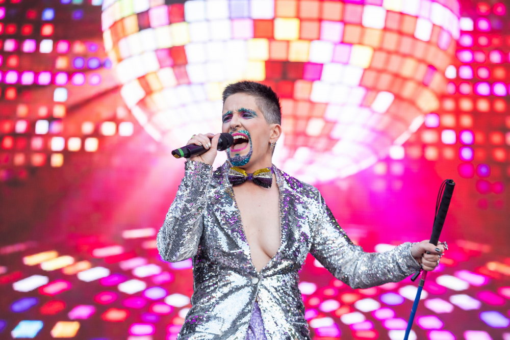 Tito Bone wears a silver sequin sparkly outfit and a glitter beard in front of a large pink glittery disco ball, singing into a microphone.