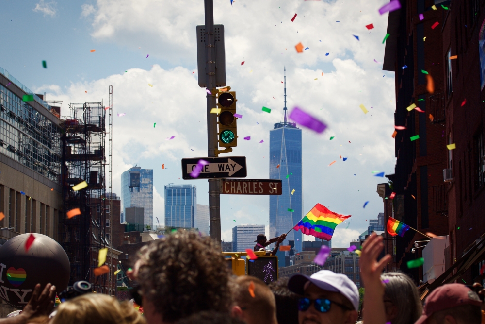 a crowd of people at New York City pride, many people are holding small rainbow flags and the new York city skyline is in the distance