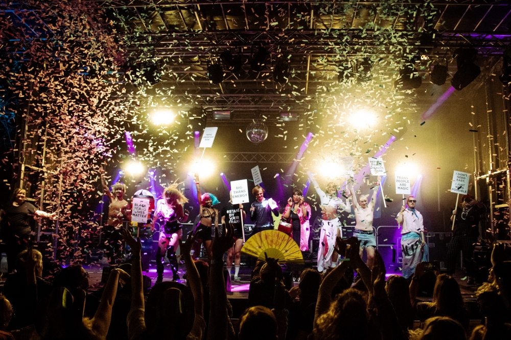 the gay village party at Manchester Pride at the peak of excitement, the crowds are jumping and yellow confetti is falling from the sky, the group on stage are dancing and holding signs which read 'big trans energy'