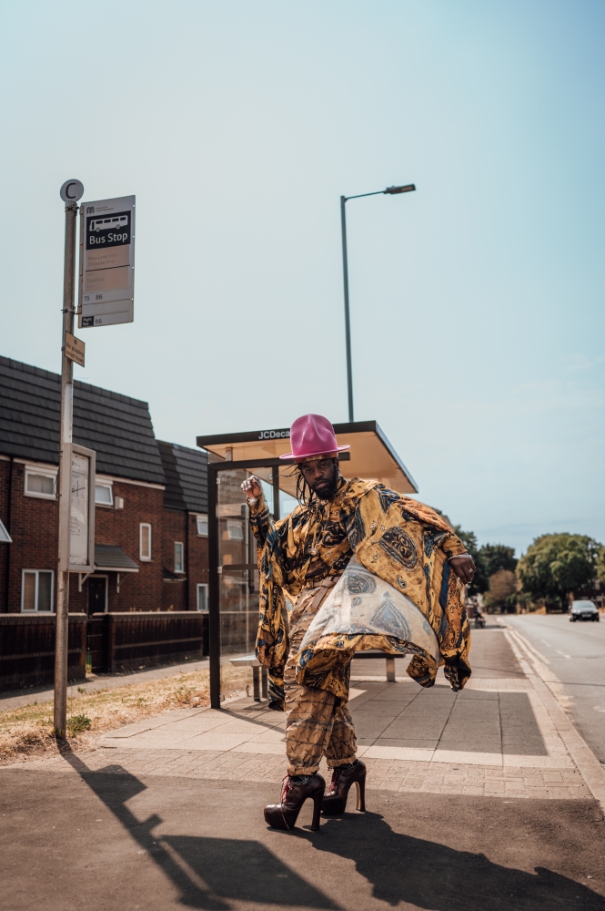 A person stands in front of a bus stop, they have dark skin, a dark beard with dark hair, a pink hat and a golden poncho, and they are wearing high platform heels.