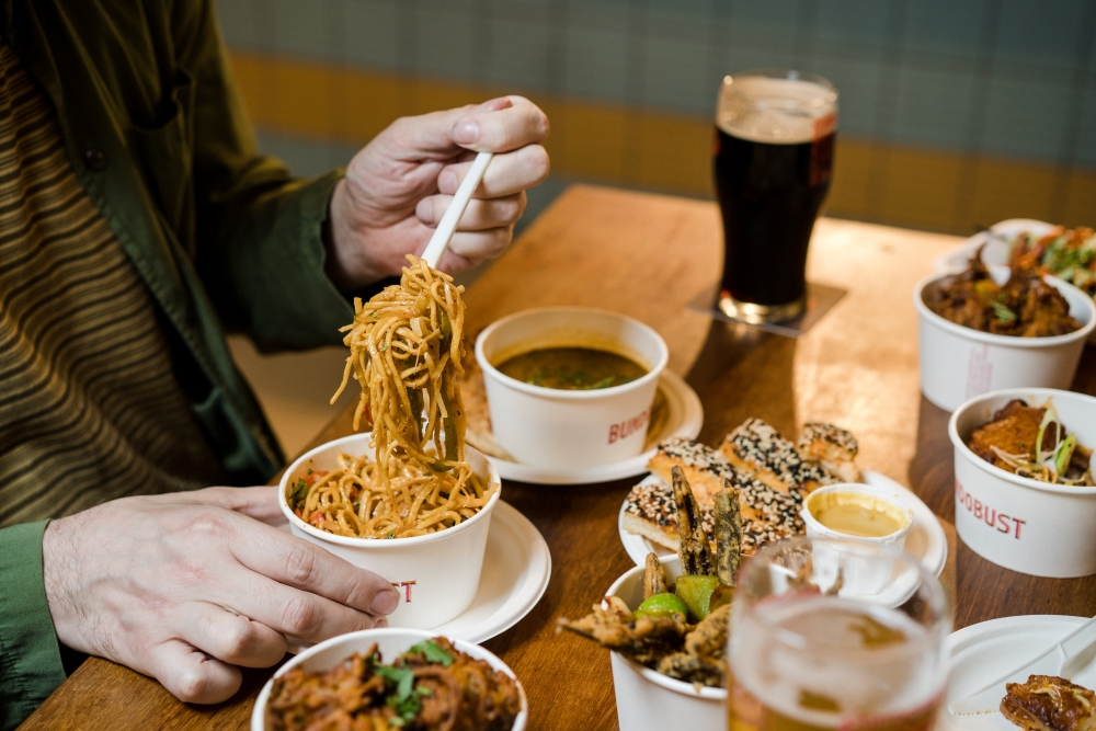 A table full of the iconic small, white, round bundobust containers filled with every dish on the new Indochinese menu. A person with white skin is digging into the Hakka noodes.