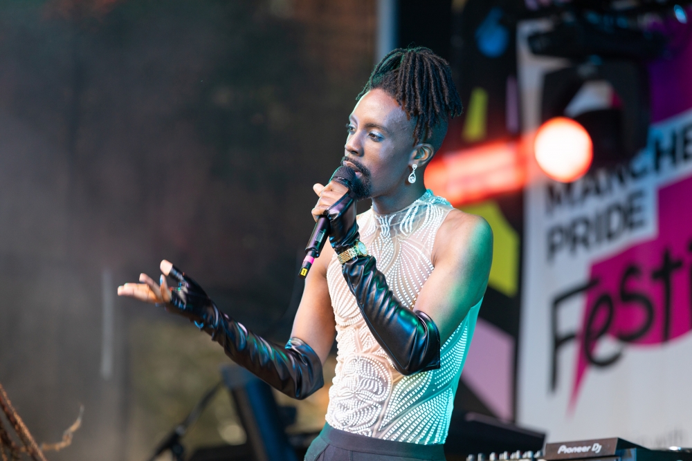 Sky, a black person with black, mid length hair tied up into a bun, wears a white vest with floral patterns and holds a microphone