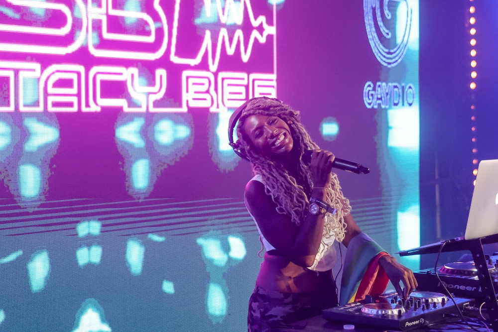 Dj Stacy Bee stop din front a big LED screen with her name and branding on it, Stacy has light brown long dreads and over-ear headphones on