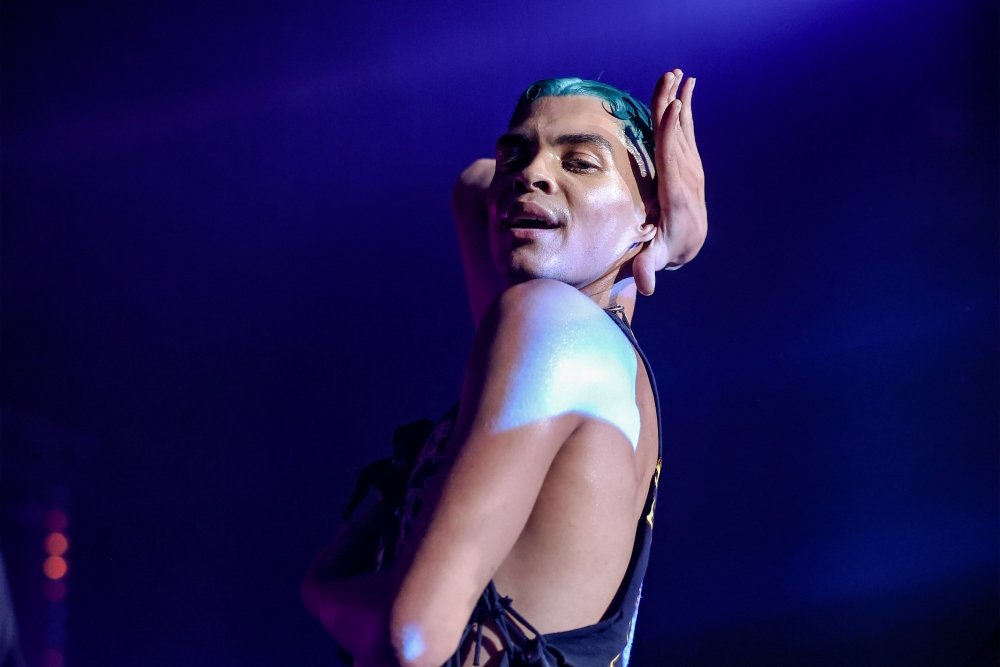 Reece is a mixed race, light brown skinned dancer with bright blue, short slicked down hair. Reece is mid vogue with their hands wrapped around their chest and head, looking directly at the camera