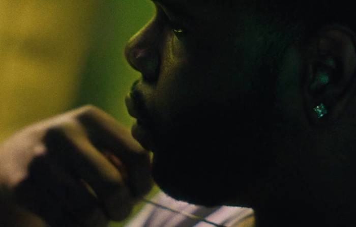 A black person with short hair, a dark beard and diamond earring looks on, inquisitively