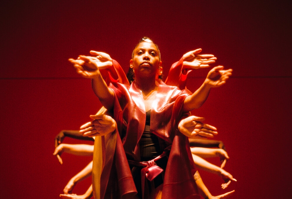 A person is dancing, looking above the camera, as several pairs of arms protrude from the people stood behind her