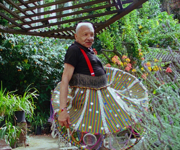 A man with a grey beard stands in a garden wearing a skirt, humurous expression on his face